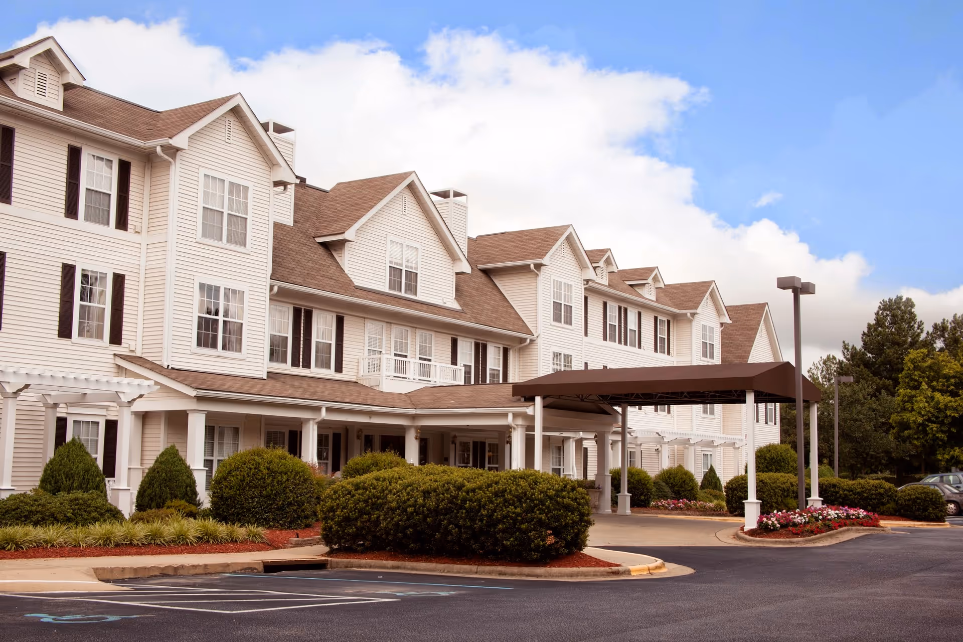 Front exterior of a multi-story senior living facility with a covered entrance, landscaped shrubs, and a parking area.