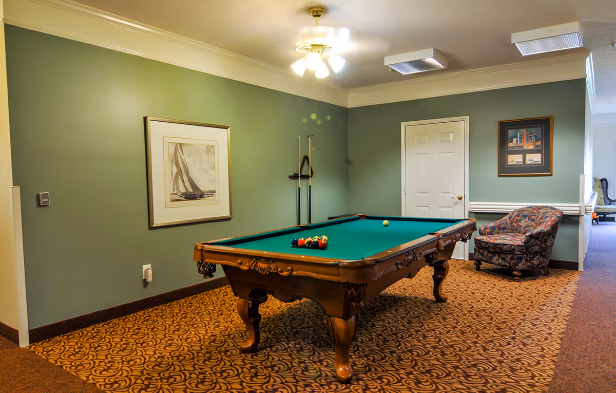 A room with a green pool table in the center, a patterned carpet, green walls with framed pictures, a ceiling fan with lights, and a cushioned armchair in the corner.