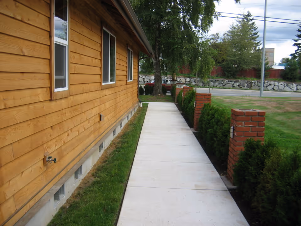 Side view of a wooden building with three windows and a concrete walkway bordered by small green bushes and brick pillars leading to a tree and a road in the background.