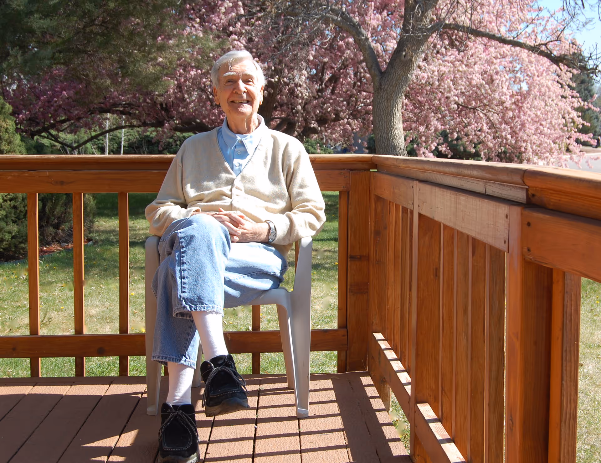 An elderly man sitting cross‑legged on a chair on a wooden deck with pink flowering trees in the background.