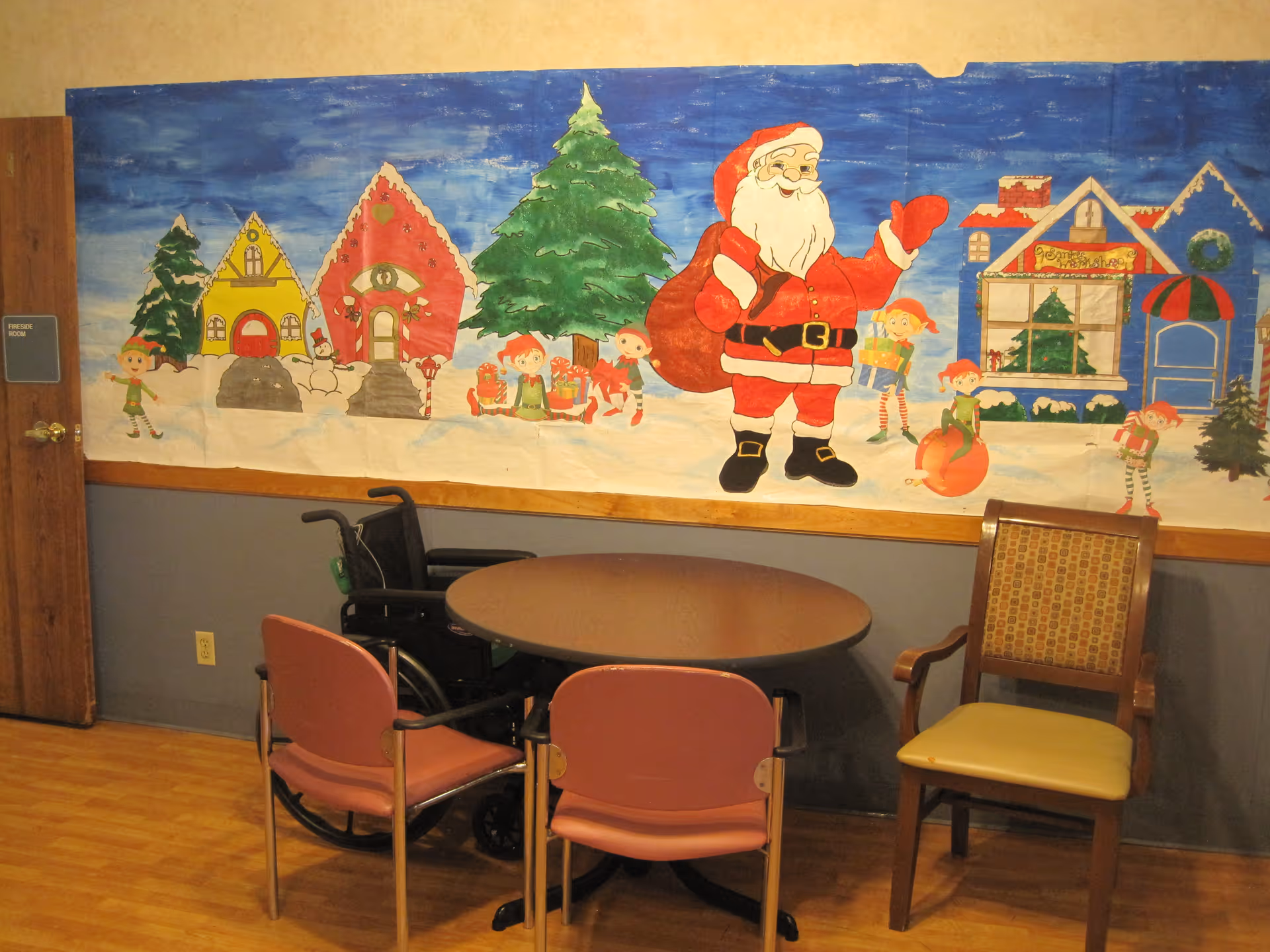 A small round table surrounded by three chairs and a wheelchair in a room with wooden flooring. On the wall behind the table is a large festive mural featuring Santa Claus, elves, Christmas trees, and colorful houses in a snowy scene.