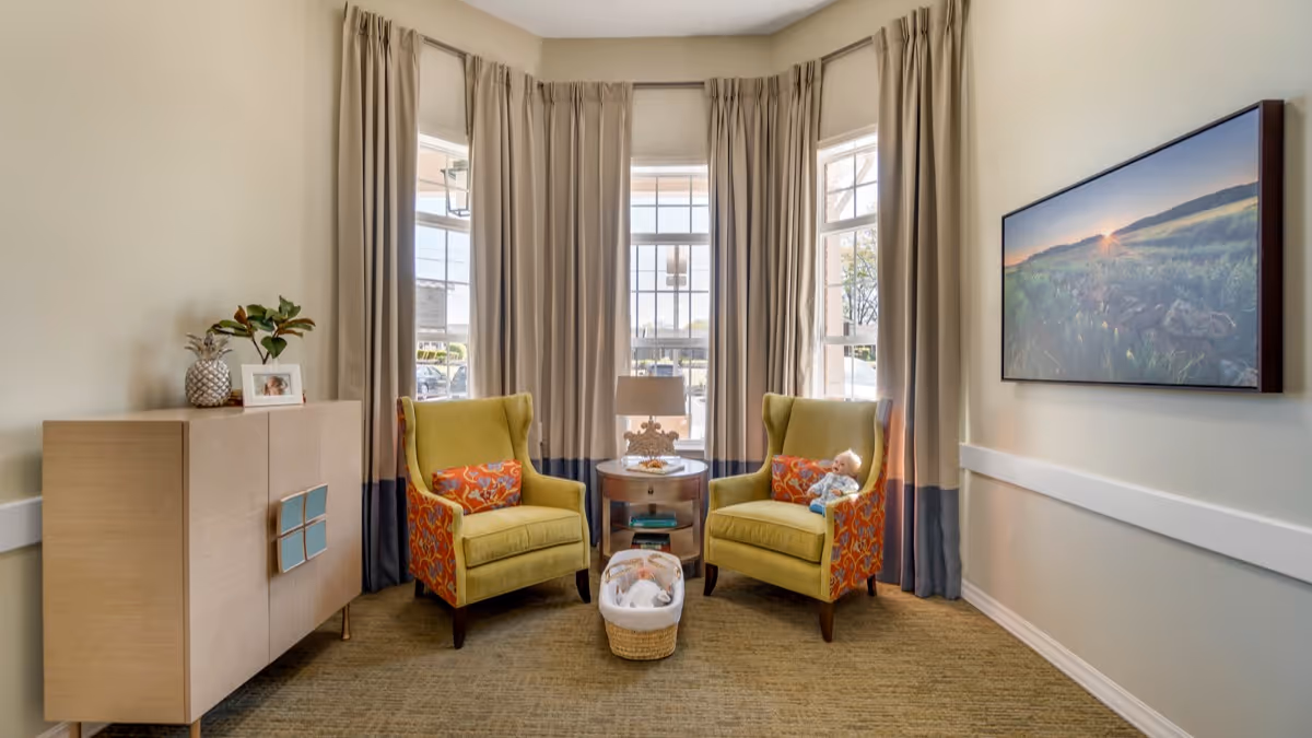 A cozy sitting area with two yellow armchairs featuring orange patterned sides and matching pillows, positioned around a small round wooden table with a lamp. A basket with a doll is placed on the carpeted floor between the chairs. Behind the chairs are three tall windows with beige and blue curtains. To the left is a light wood cabinet with decorative items including a pineapple and a framed photo. On the right wall hangs a large landscape photograph of a sunrise over a field.