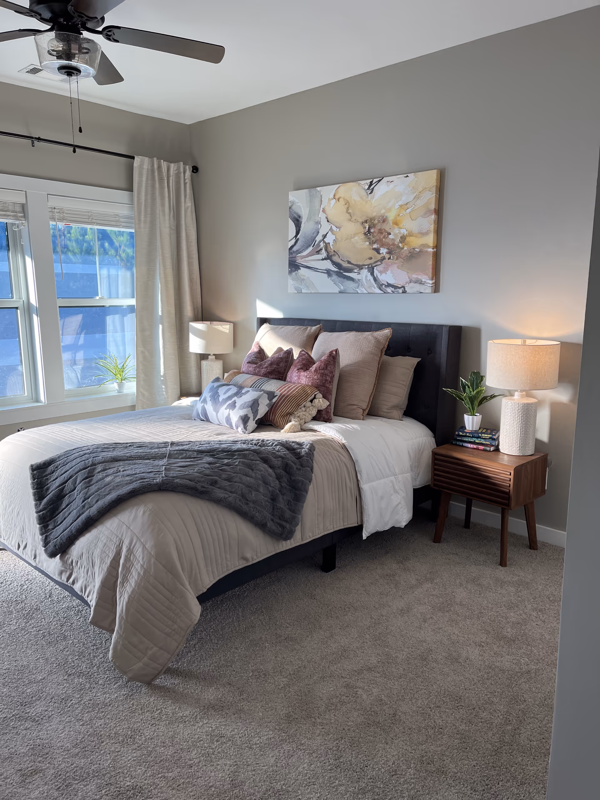 A neatly made bed in a bedroom with beige and gray bedding, multiple pillows including purple and patterned ones, and a gray throw blanket. The room has light gray walls, a ceiling fan, a large window with beige curtains, a wooden nightstand with a lamp, a small plant, and some books. Above the bed hangs an abstract painting with soft colors.