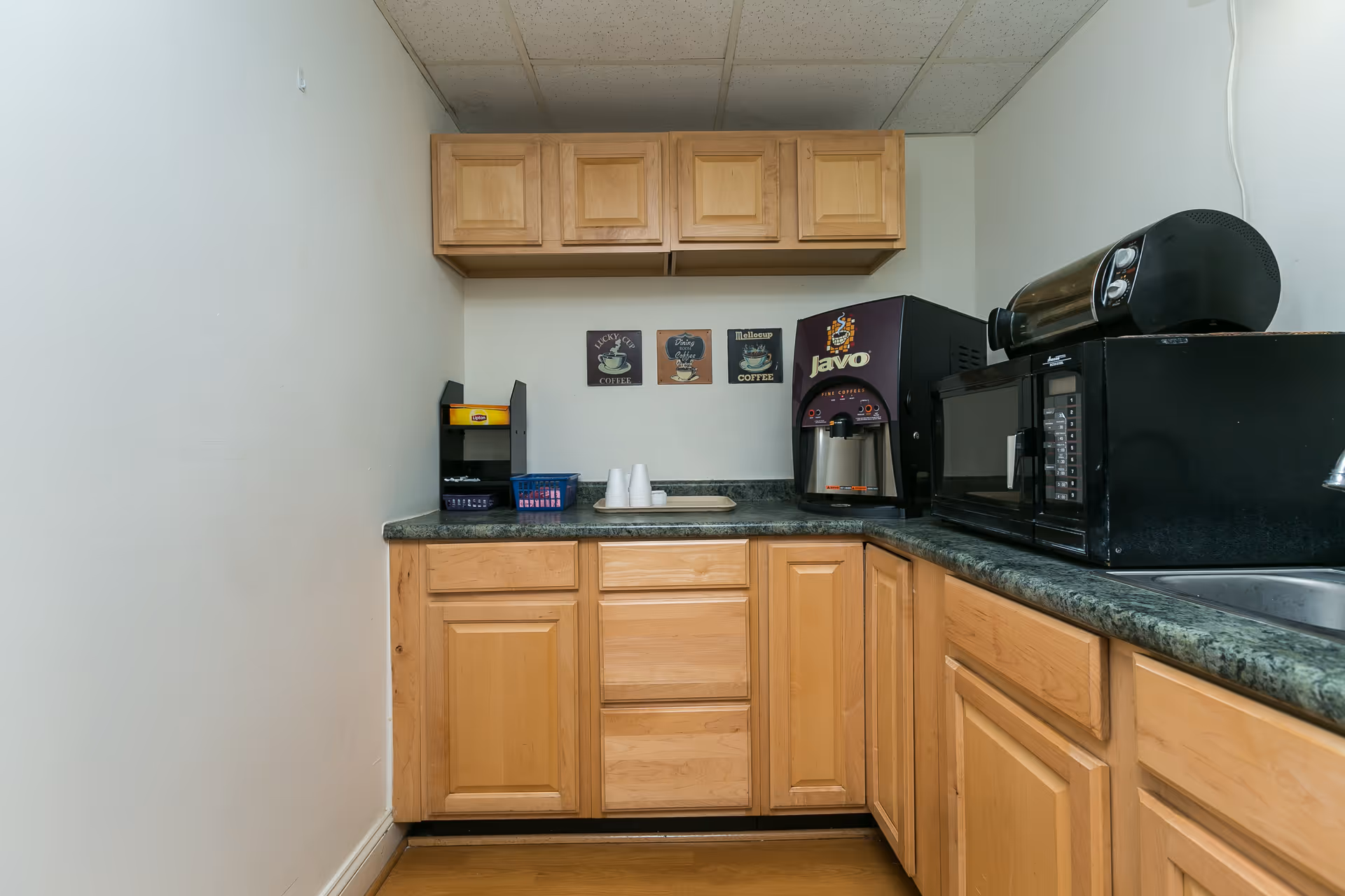 Small kitchenette area with wooden cabinets, a coffee machine, a microwave, a toaster oven, a sink, and a countertop with disposable cups and a tray. Three small coffee-themed decorative signs are mounted on the wall above the counter.