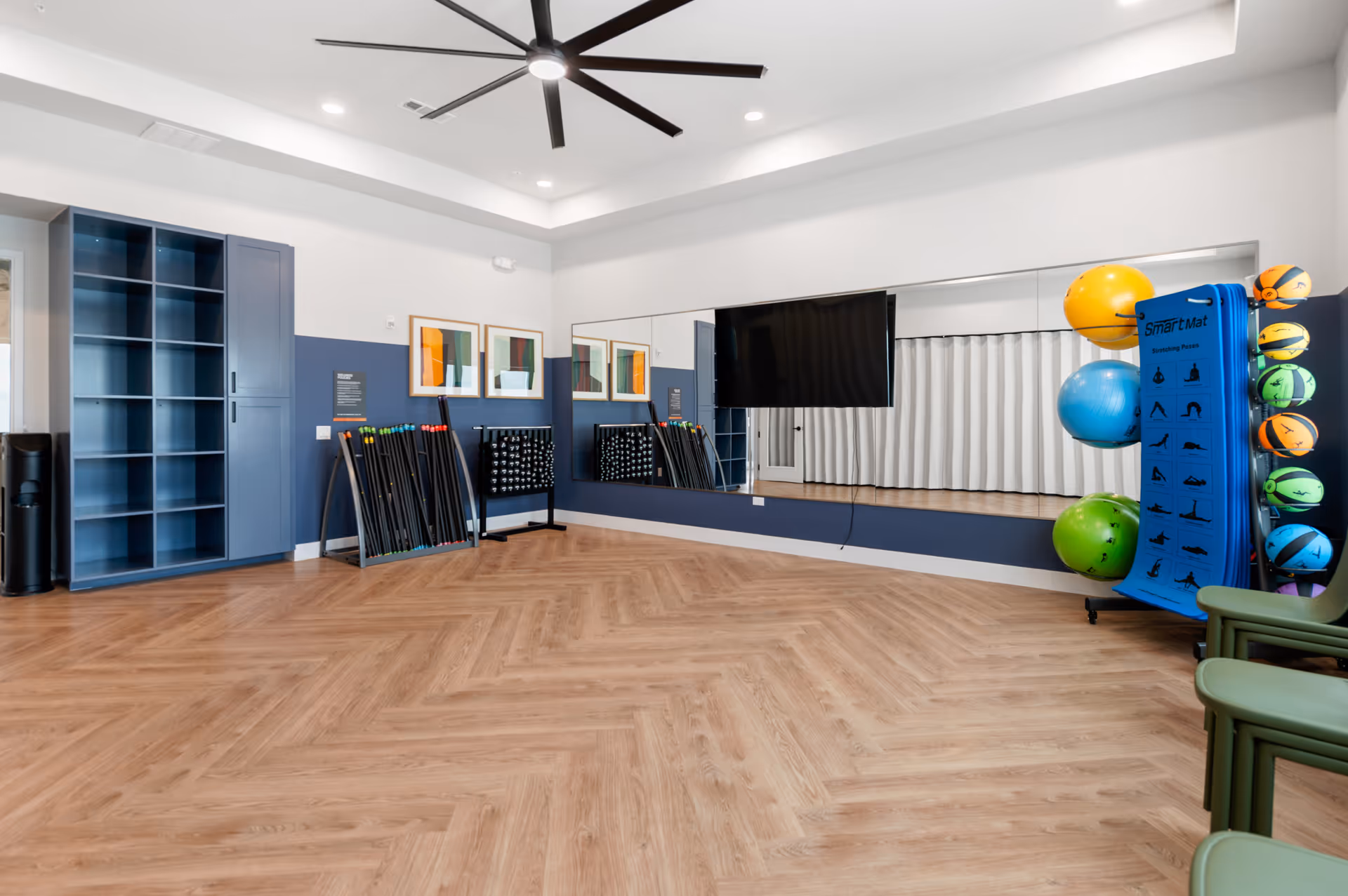 Spacious interior activity/fitness room with herringbone wood floor, wall mirror, mounted TV, storage shelves and exercise balls.