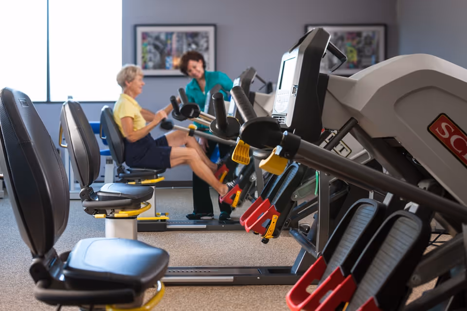 An elderly woman exercises on a seated leg press machine in a fitness room while a caregiver or trainer assists her. The room has multiple exercise machines and a window letting in natural light.
