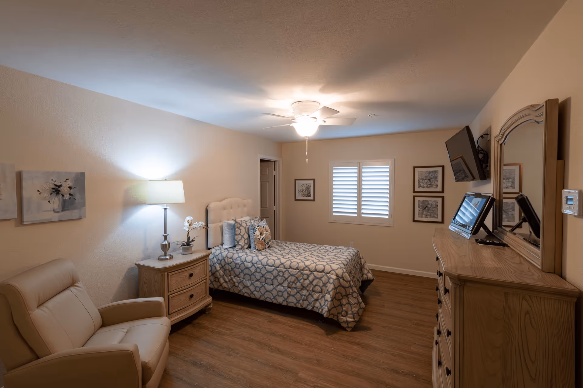 A cozy bedroom with a single bed covered in a patterned blue and beige bedspread. Next to the bed is a wooden nightstand with a lamp and a small plant. There is a beige armchair in the corner, wooden flooring, and a ceiling fan with a light. On the opposite wall, there is a wooden dresser with a mirror and a mounted flat-screen TV. The room has beige walls with framed artwork and a window with white shutters.