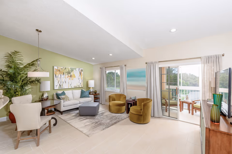 A bright and spacious living room with a white sofa, two mustard yellow armchairs, a gray ottoman, and a wooden TV stand with a flat-screen TV. The room has light-colored tiled flooring, a green accent wall with a large painting, and large windows with beige curtains. There is a sliding glass door leading to a balcony with outdoor seating and a view of trees.