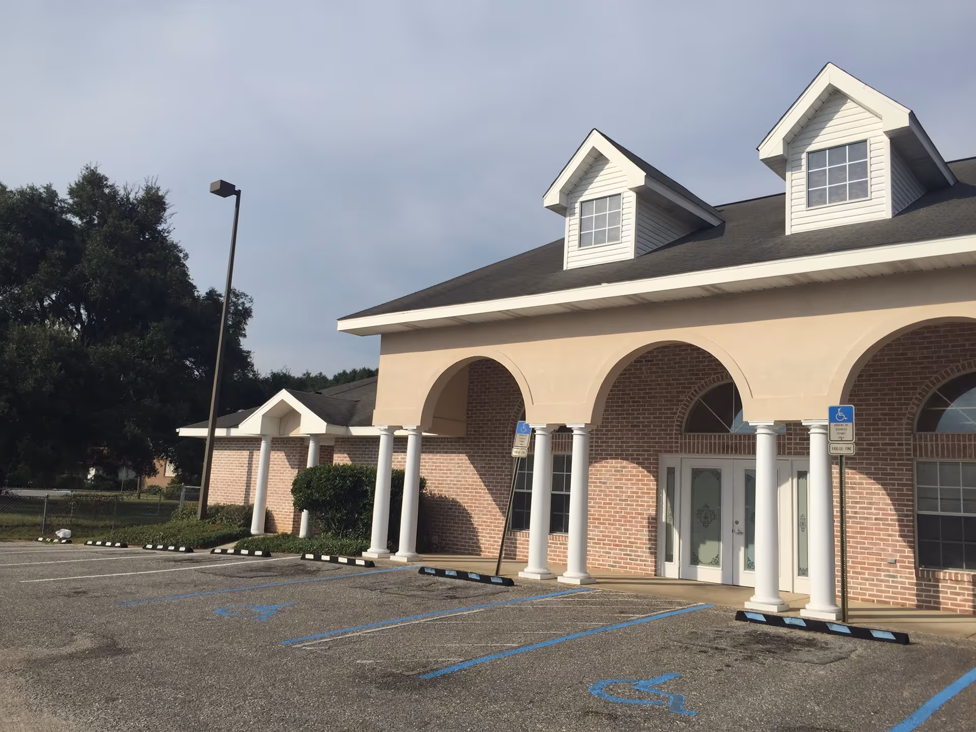 Exterior view of a brick building with white columns and arches at the entrance, featuring two dormer windows on the roof and several handicap parking spaces in front.