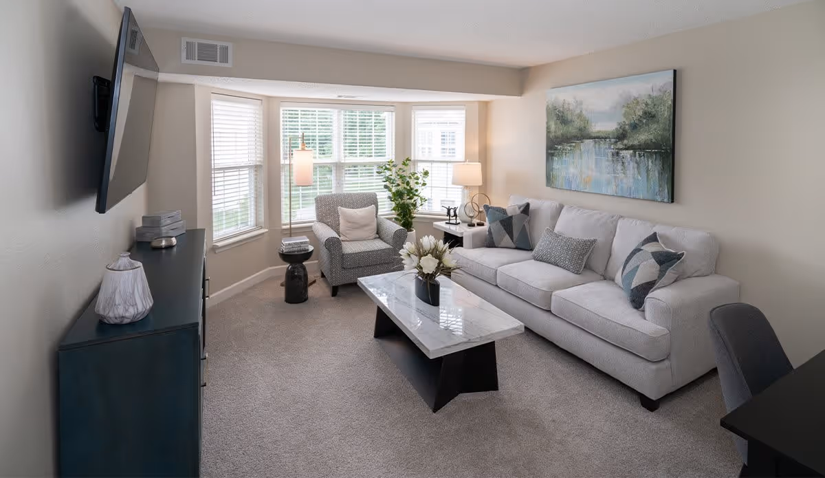 A cozy living room with a light gray sofa adorned with patterned pillows, a matching armchair with a white cushion, a marble coffee table with a flower arrangement, a side table with a lamp and decorative items, a large window with white blinds, a wall-mounted flat-screen TV, and a landscape painting above the sofa.