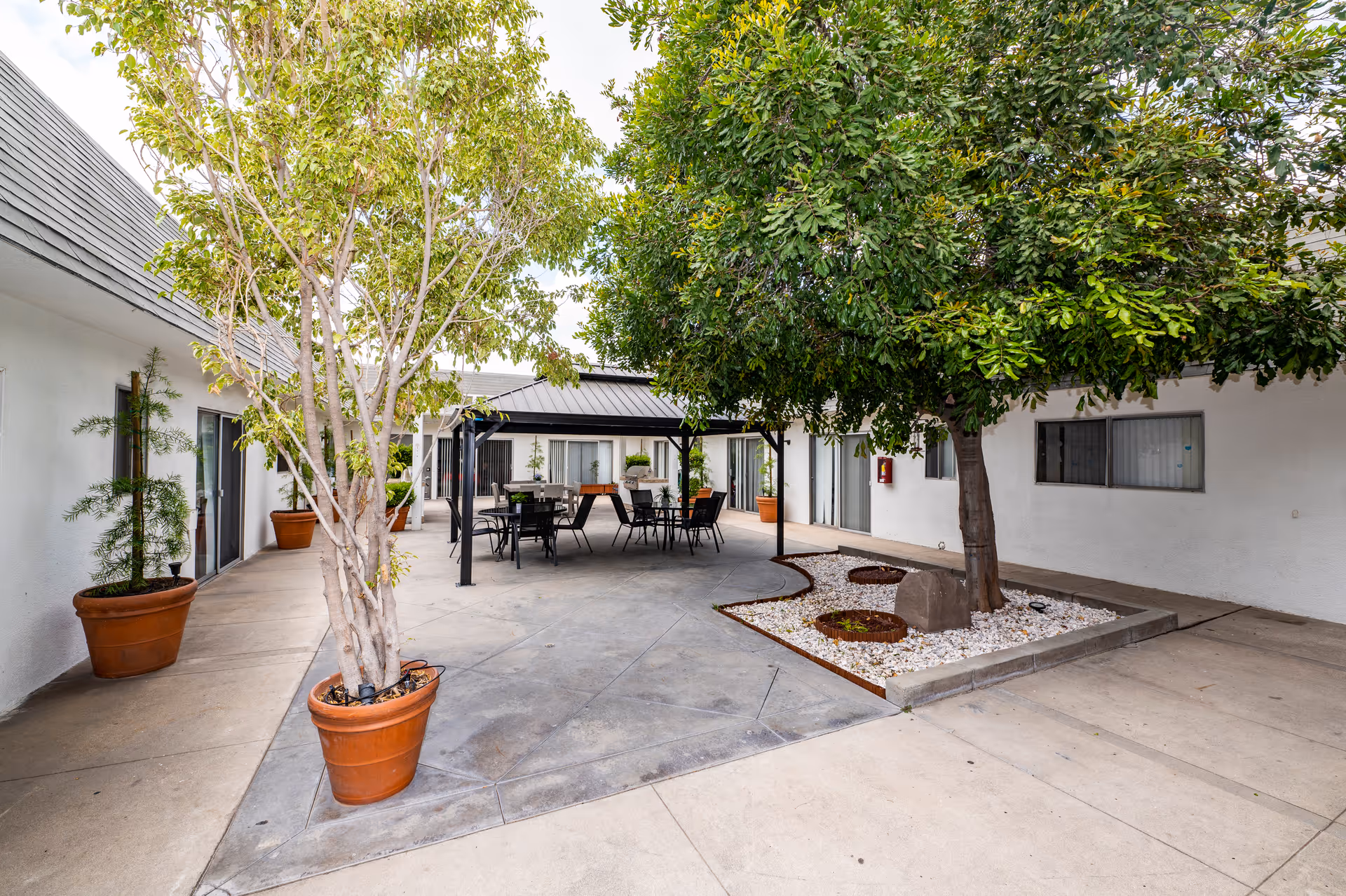 Outdoor courtyard area at Citrus Nursing Center featuring potted trees, a large tree planted in a white rock bed, a shaded seating area with tables and chairs, and white building walls with windows and sliding doors surrounding the space.