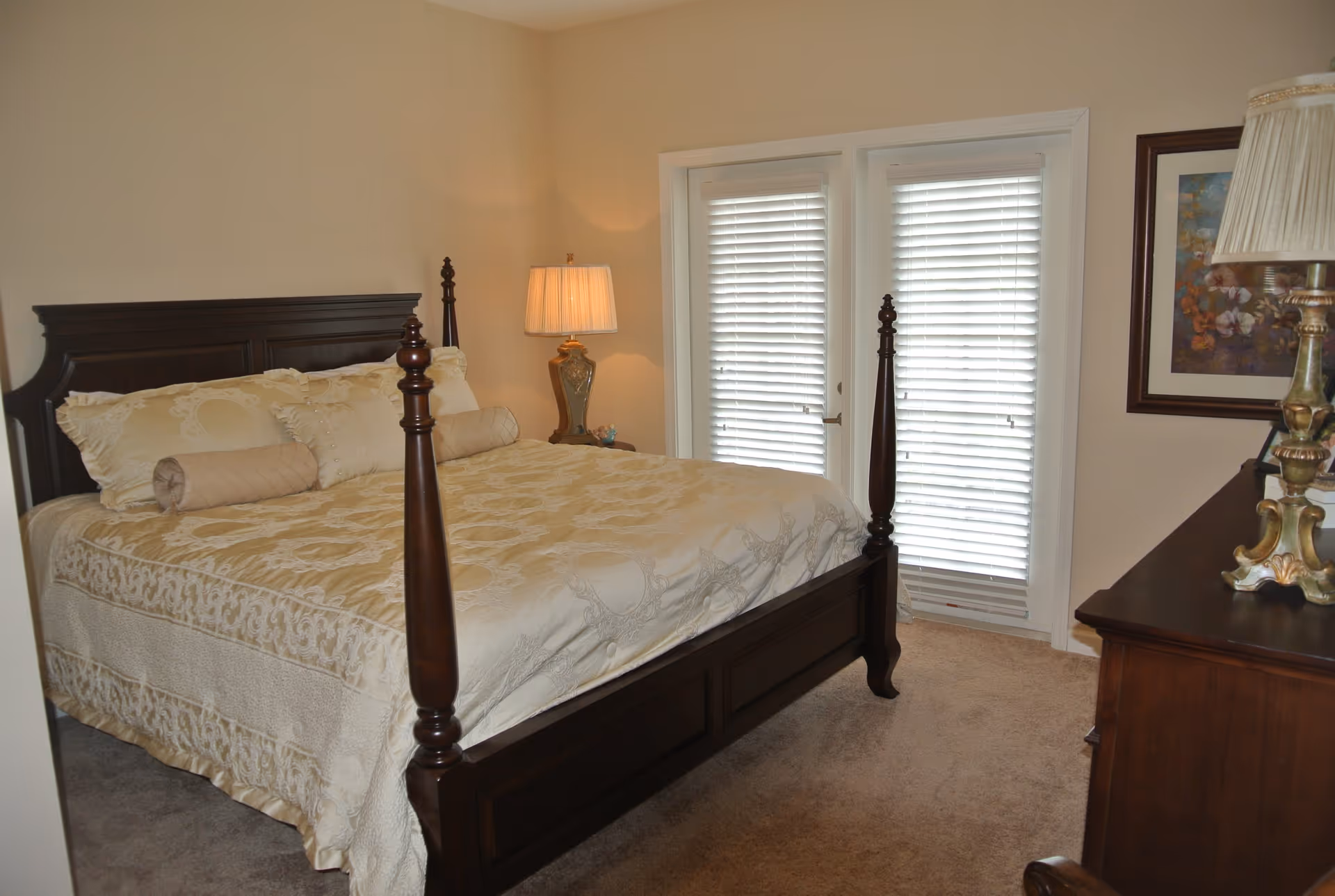 A bedroom with a large dark wooden four-poster bed dressed in beige and cream bedding. There is a nightstand with a lamp on the left side of the bed, and a dresser with a decorative lamp and framed artwork on the right. Two glass doors with white blinds are visible behind the bed.