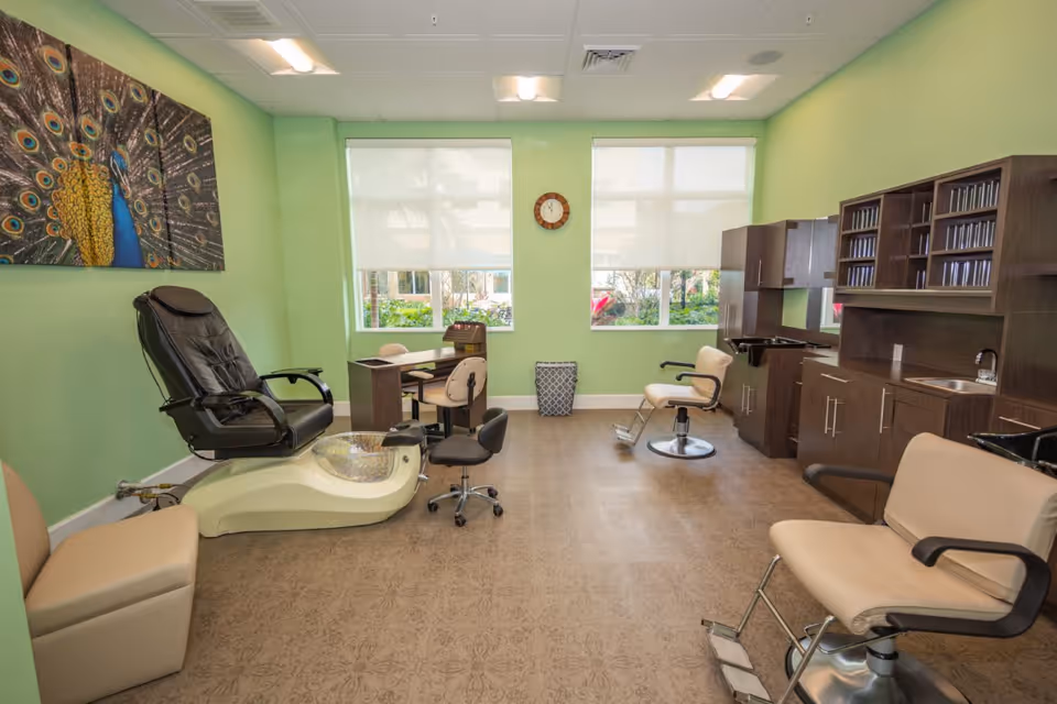 Interior of a salon room with light green walls, featuring a pedicure chair with a foot bath, two salon chairs with footrests, a desk with two chairs, and wooden cabinetry with a sink and shelves. A peacock painting is hung on the left wall, and two large windows with white blinds allow natural light into the room.