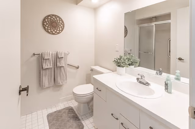 A clean and bright bathroom featuring a white toilet, a white countertop with a sink, a large mirror above the sink, a potted plant, soap dispensers, a towel rack with two beige towels, a decorative wall piece, a gray bath mat on the tiled floor, and a glass-enclosed shower in the background.
