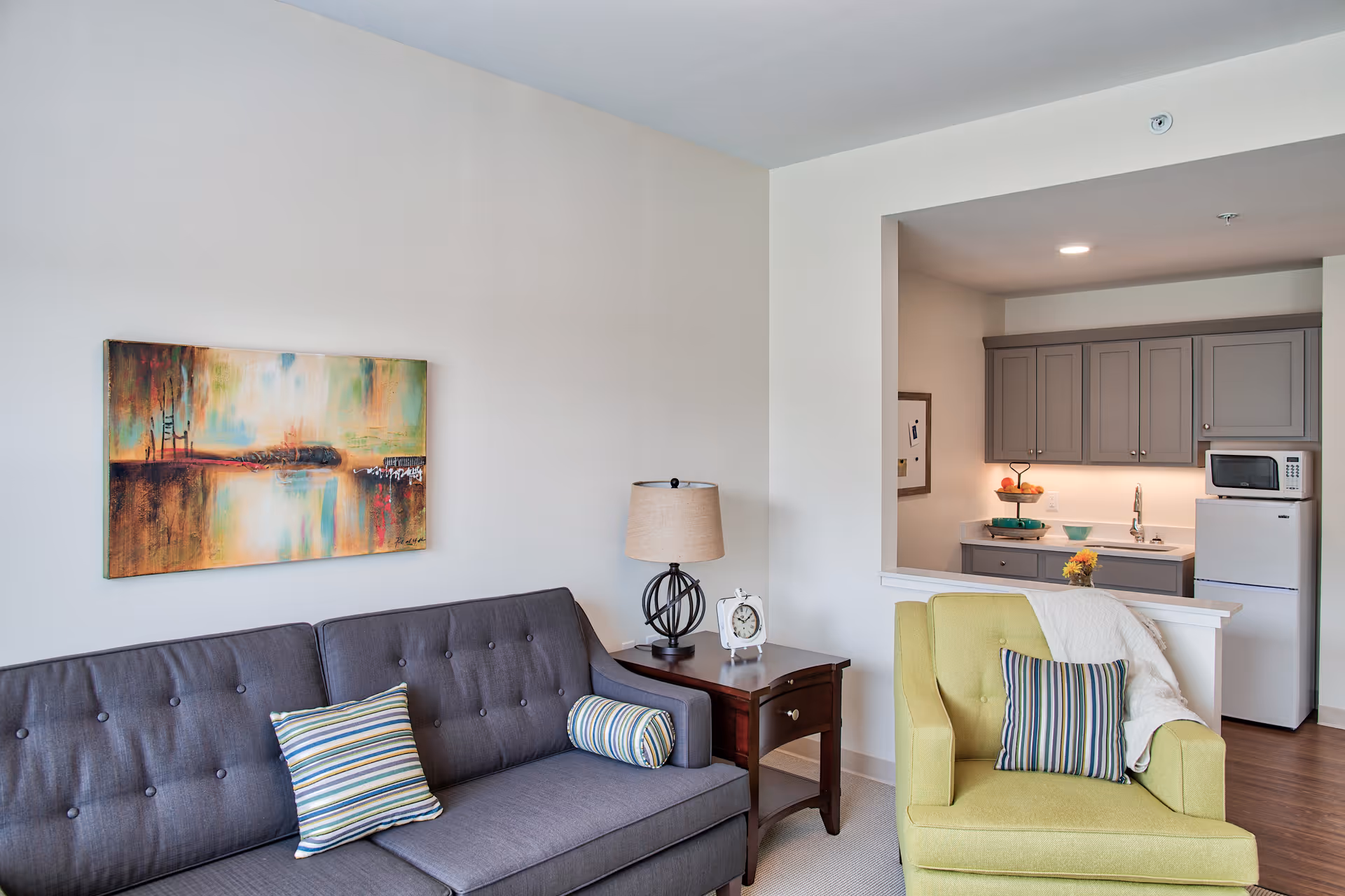 A cozy living room area featuring a dark gray tufted sofa with striped pillows, a wooden side table with a lamp and a clock, and a light green armchair with a striped pillow and a white throw blanket. In the background, there is a small kitchen area with gray cabinets, a white refrigerator, a microwave, and a countertop with a fruit basket and a small vase of flowers. A colorful abstract painting hangs on the wall above the sofa.