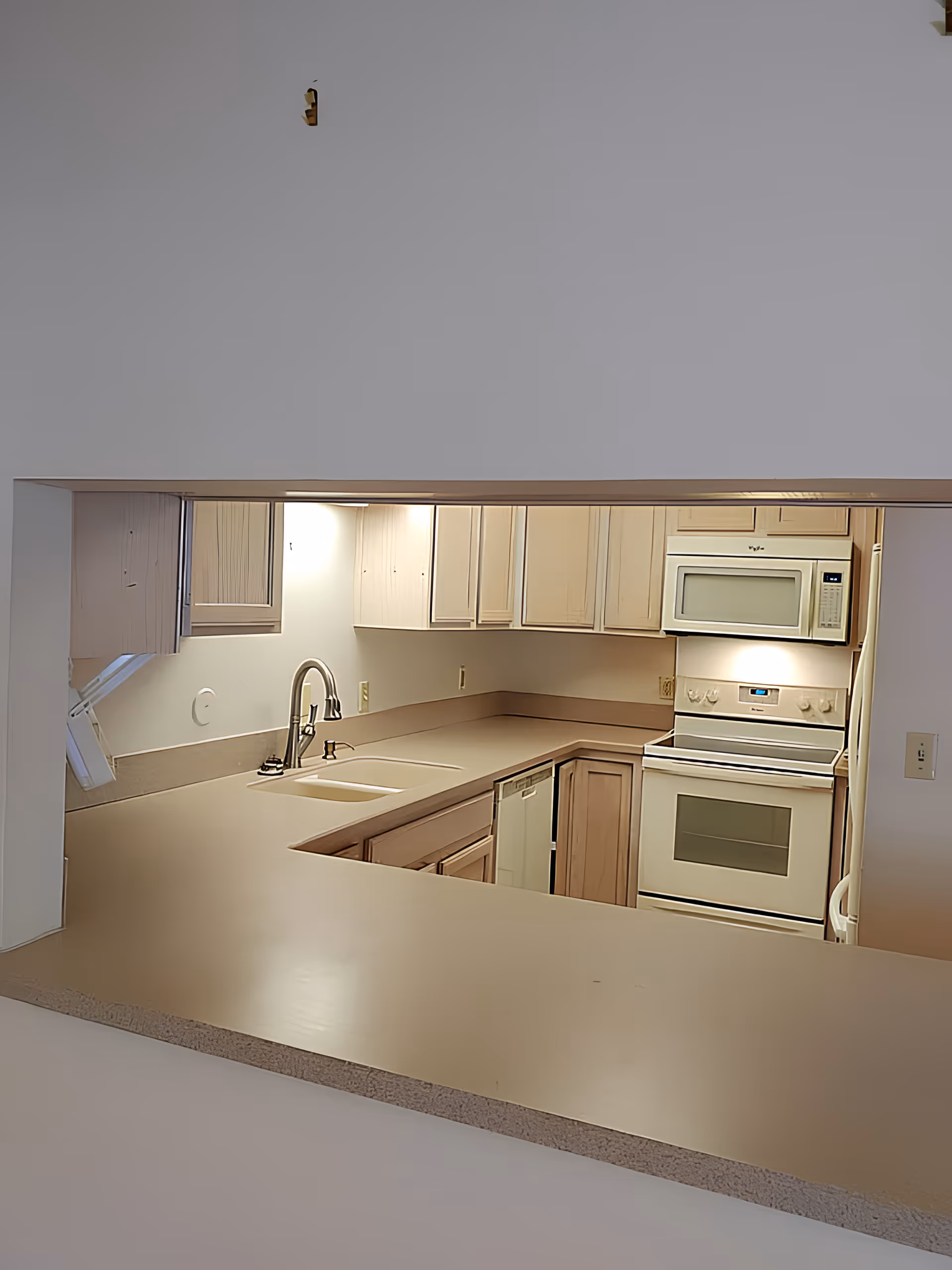 U-shaped kitchen with beige countertops, light wood cabinets, a sink and faucet, dishwasher, stove and microwave visible through a pass-through opening.