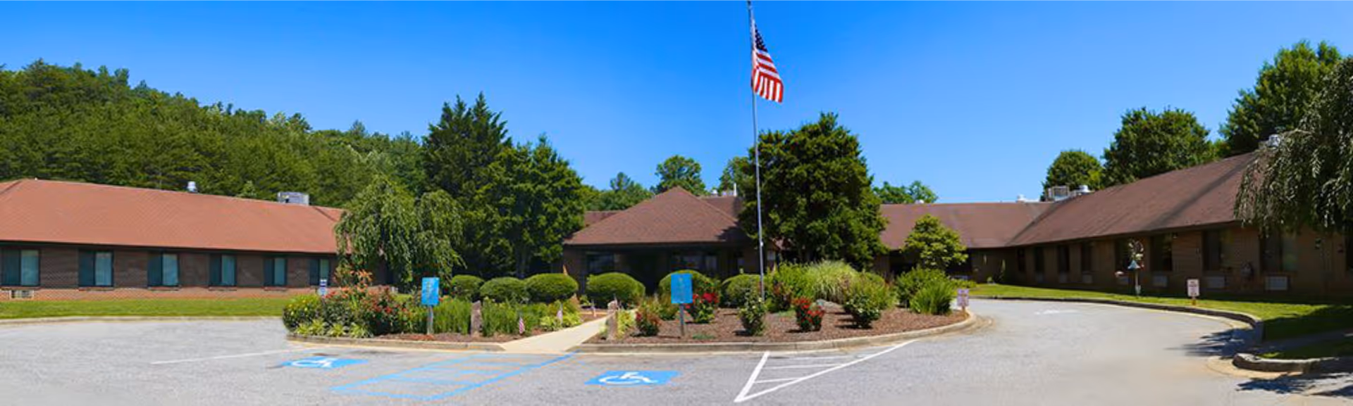 Wide exterior view of Chatuge Regional Nursing Home showing a U-shaped single-story brick building with a brown roof, surrounded by landscaped greenery and trees. An American flag is prominently displayed on a flagpole in the center of a circular garden bed. Several handicap parking spaces are visible in the foreground under a clear blue sky.