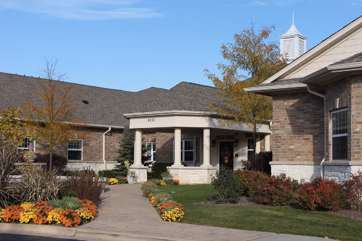 Front entrance of a brick senior living facility with a columned portico, walkway and fall flower beds.