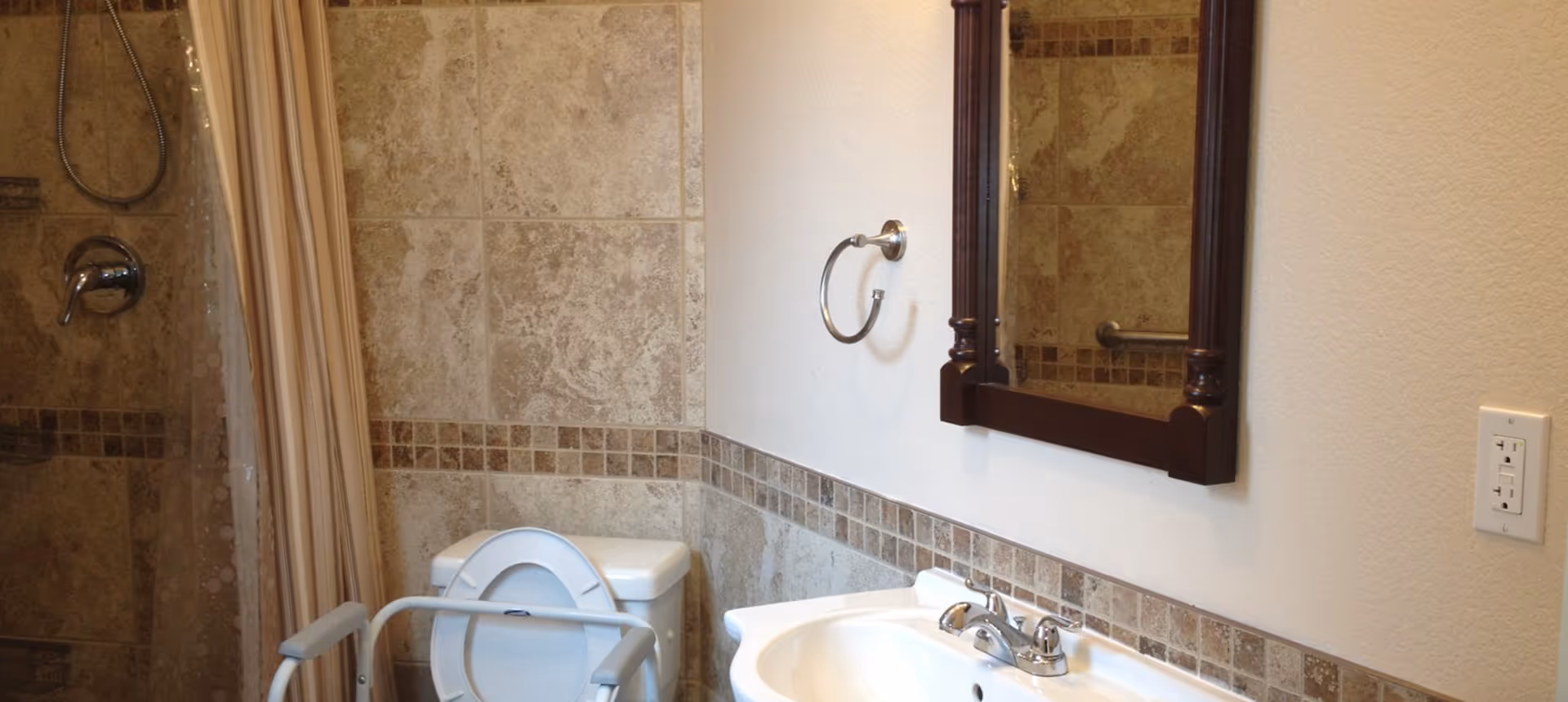 A bathroom with beige tiled walls, a white toilet with a support frame, a white sink with a chrome faucet, a wooden framed mirror above the sink, a towel ring on the wall, and a shower area with a curtain and handheld showerhead.