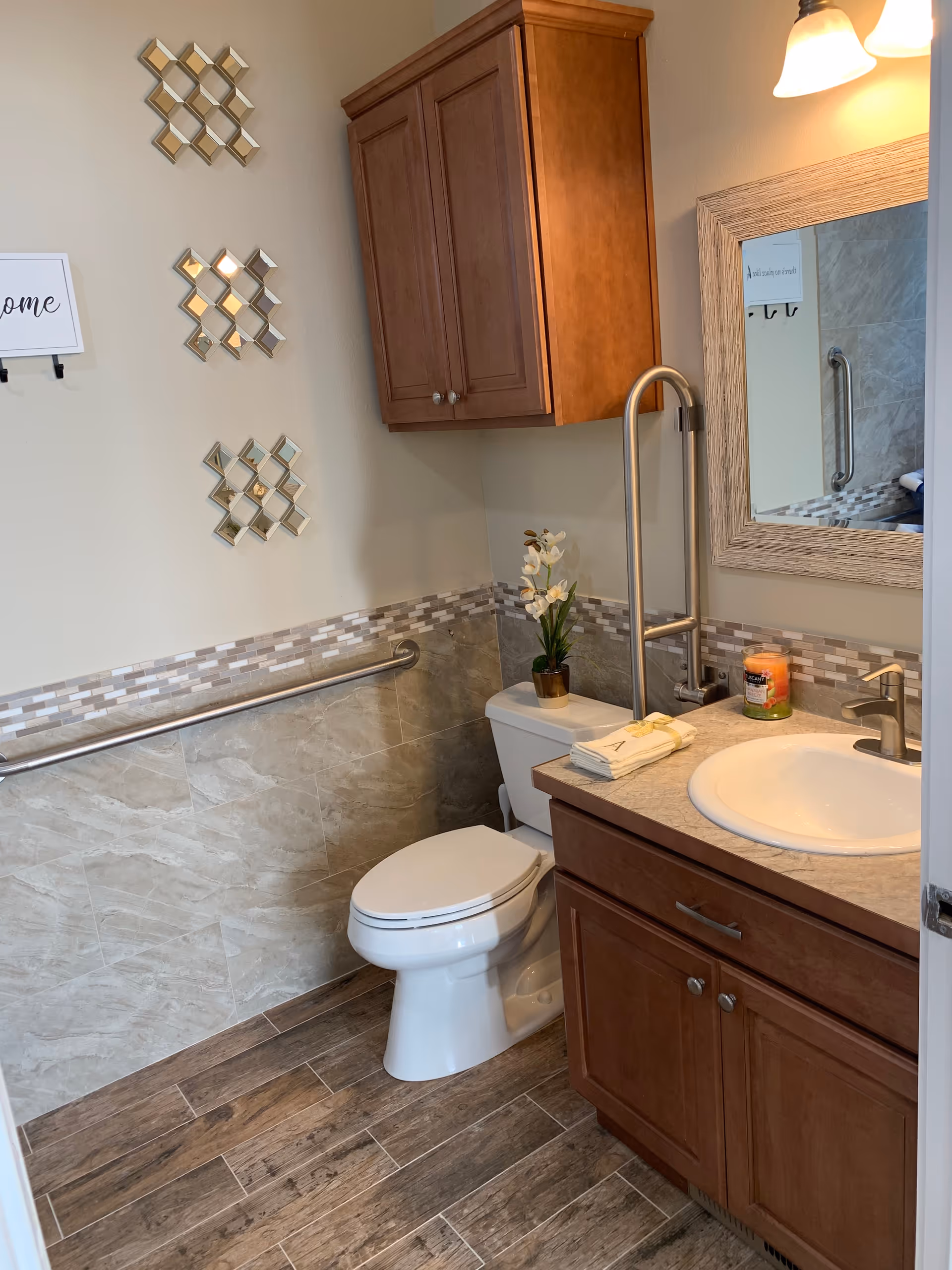 A clean and well-lit bathroom with a white toilet, a wooden cabinet above it, and a wooden vanity with a sink and faucet. There is a large mirror above the sink, a candle, and folded towels on the countertop. The walls have a decorative tile border and a grab bar is installed next to the toilet. The floor has wood-like tiles and decorative wall art is visible on the left wall.