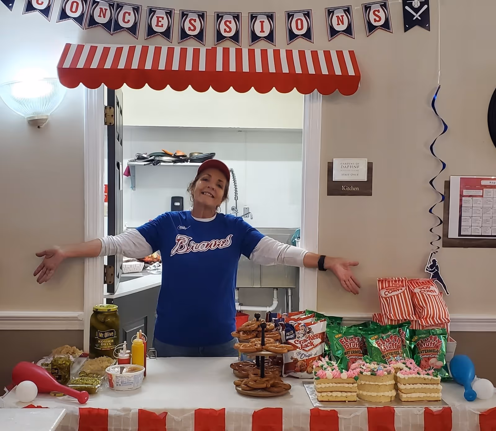 A woman wearing a blue Braves shirt and red cap stands behind a concession stand table with arms open wide. The table is decorated with a red and white striped tablecloth and has various snacks including pretzels, popcorn bags, chips, pickles, and small cakes. Above her is a red and white striped awning with a banner spelling 'CONCESSIONS'. The setting appears to be indoors with a kitchen visible behind her.