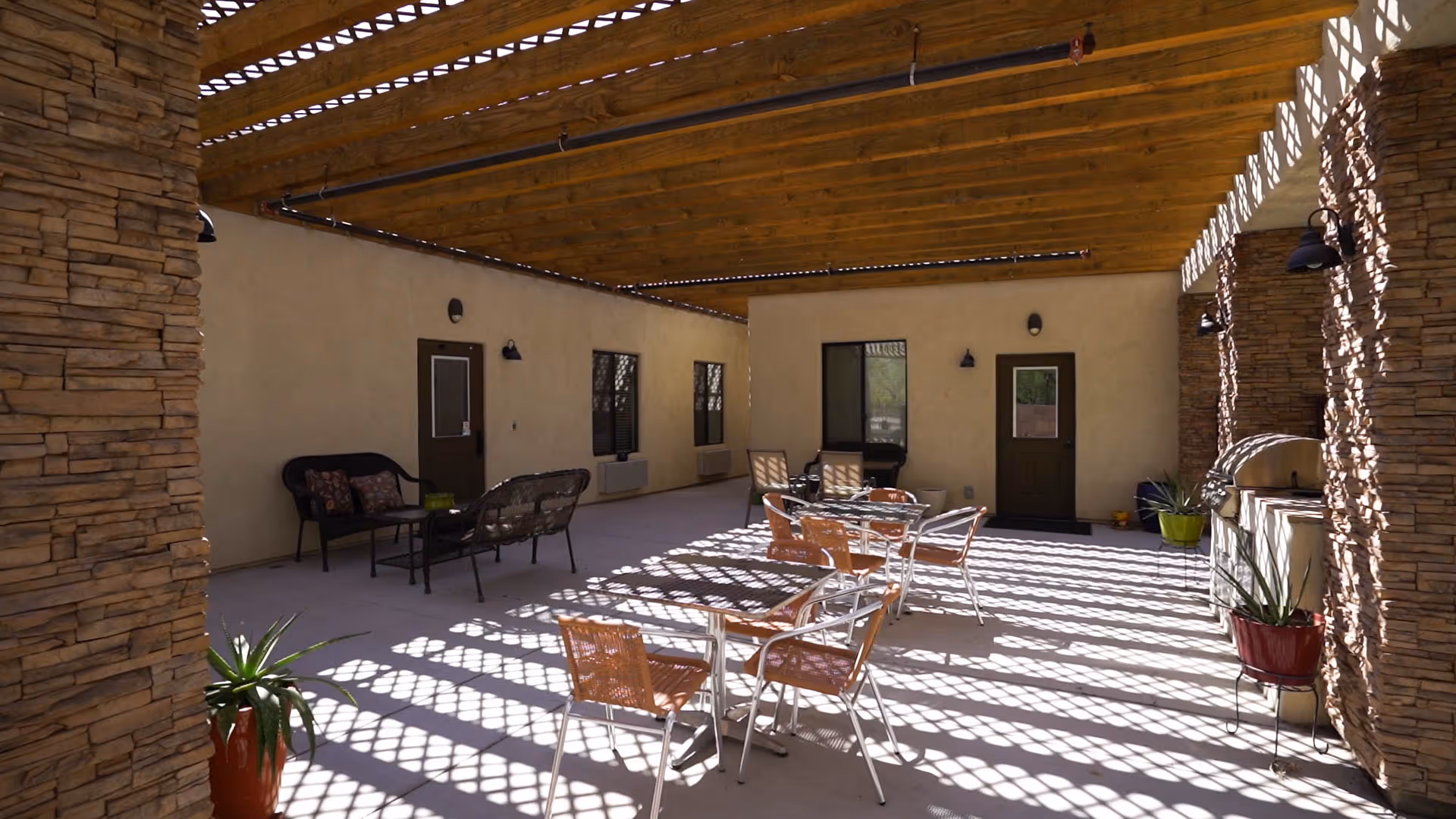 Covered outdoor patio area with wooden beams casting patterned shadows on the floor. The space includes several tables with chairs, a seating area with a loveseat and chair, potted plants, and a built-in grill. The walls are beige with dark brown doors and windows.