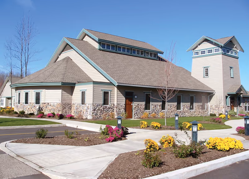 Front exterior of a single-story stone-and-siding community building with a small tower, landscaped flowerbeds, and paved walkways under a clear blue sky.