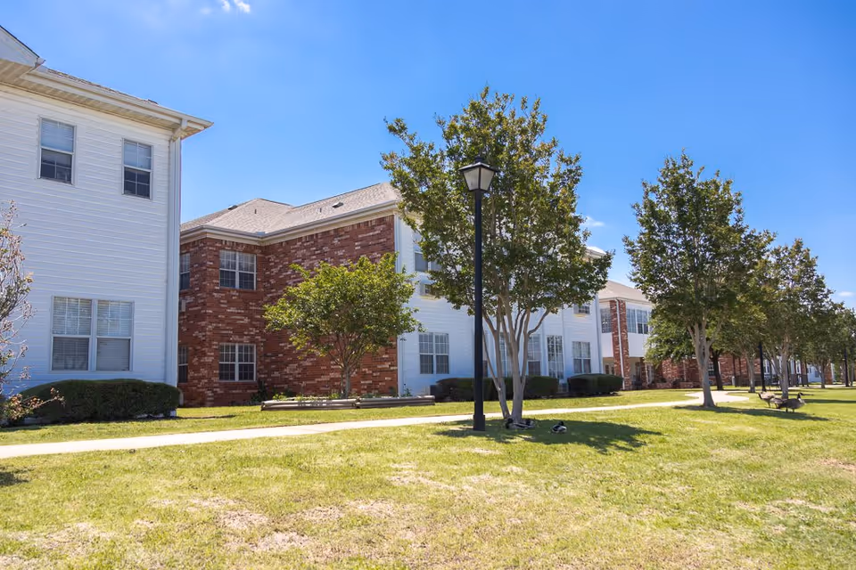 Exterior view of a senior living facility building with a combination of white siding and red brick walls. The building is two stories tall with multiple windows. There is a well-maintained grassy area in front with several trees, a sidewalk, benches, and a black lamp post under a clear blue sky.