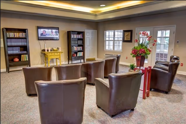 A cozy living room area with six brown leather chairs arranged in two rows facing a wall-mounted TV. The room has two tall bookshelves filled with books, a small yellow table with items on it, a window, a door, and a decorative flower arrangement on a red stand. The ceiling has recessed lighting with a warm glow.