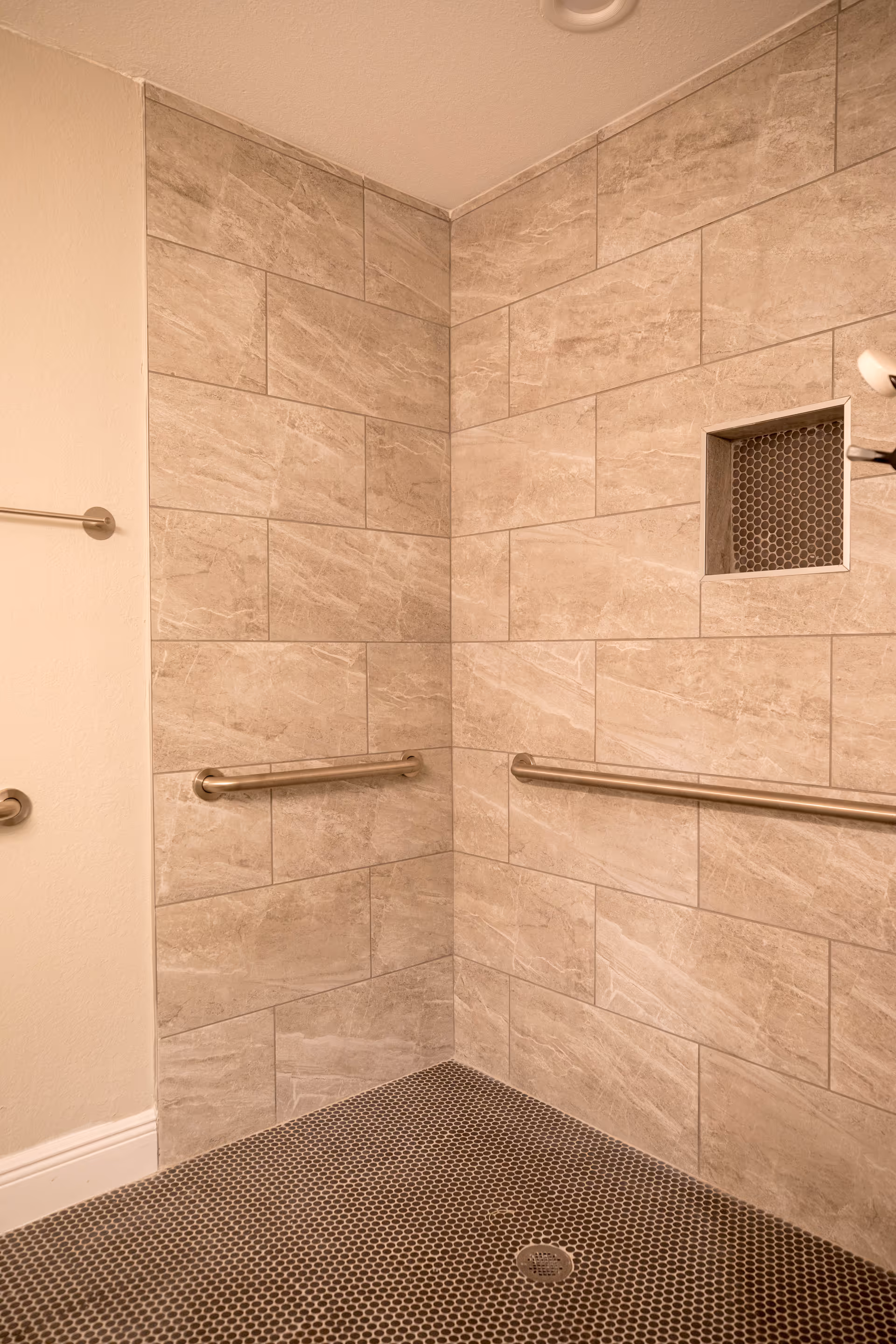 A corner of a shower area with beige stone tiles on the walls and small dark hexagonal tiles on the floor. There are two horizontal stainless steel grab bars mounted on the walls and a small recessed shelf with dark hexagonal tiles inside. A towel bar is visible on the adjacent wall outside the shower area.