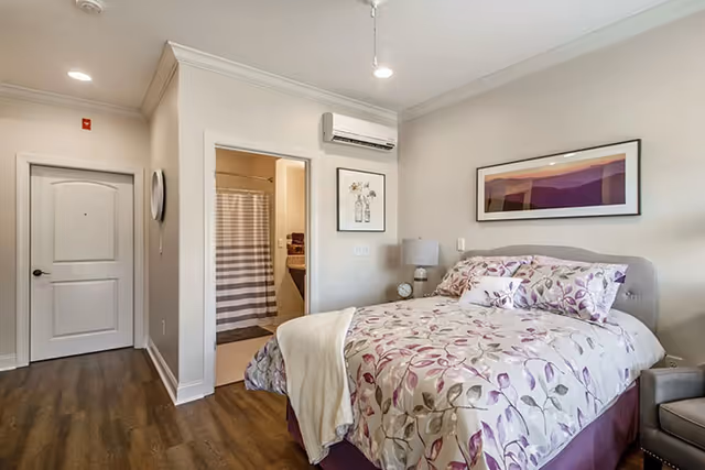 A bedroom with a bed covered in floral-patterned bedding in shades of purple and white. There is a beige blanket draped over the foot of the bed. To the right of the bed is a small nightstand with a lamp and an alarm clock. Above the bed hangs a framed landscape picture with purple tones. The room has wood flooring and light-colored walls. A door on the left leads to a bathroom with a striped shower curtain visible. Another closed door is on the left wall, and a split air conditioning unit is mounted above the bathroom door.