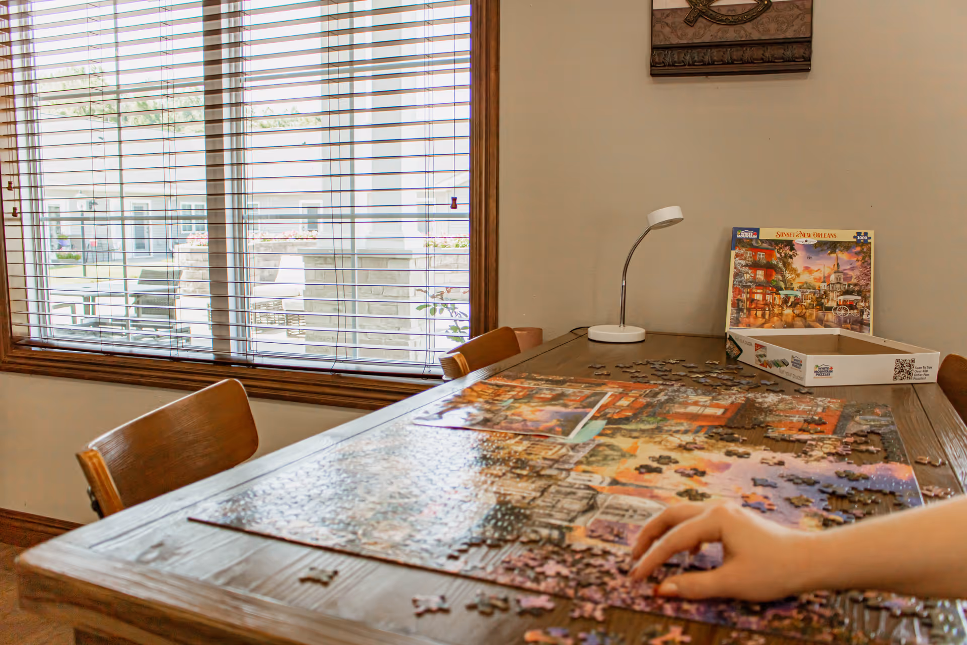 A wooden table by a window with a partially assembled jigsaw puzzle, puzzle box, lamp, and a hand placing pieces.