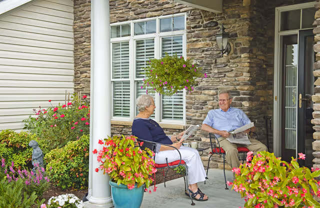 An elderly man and woman sitting on chairs on a stone porch surrounded by colorful flowers and greenery, smiling and holding newspapers, with a hanging flower basket above them and a window and door in the background.