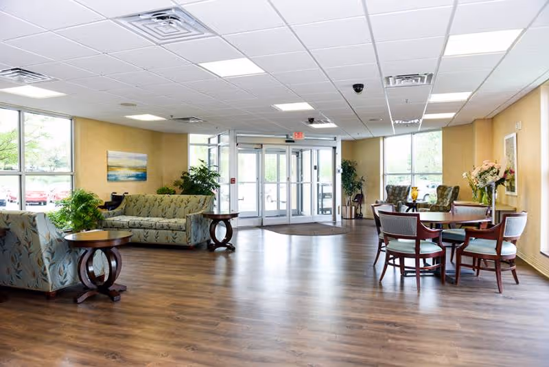 A spacious and well-lit common area with wooden flooring, featuring several seating arrangements including cushioned sofas, armchairs, and a round table with chairs. Large windows allow natural light to fill the room, and glass double doors lead outside. The walls are painted a warm yellow, decorated with framed artwork and plants.