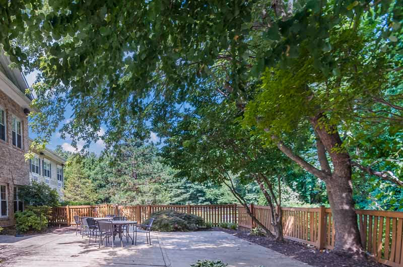 Outdoor patio area at Sterling Care Bethesda with metal tables and chairs on a concrete surface, surrounded by a wooden fence and lush green trees providing shade.