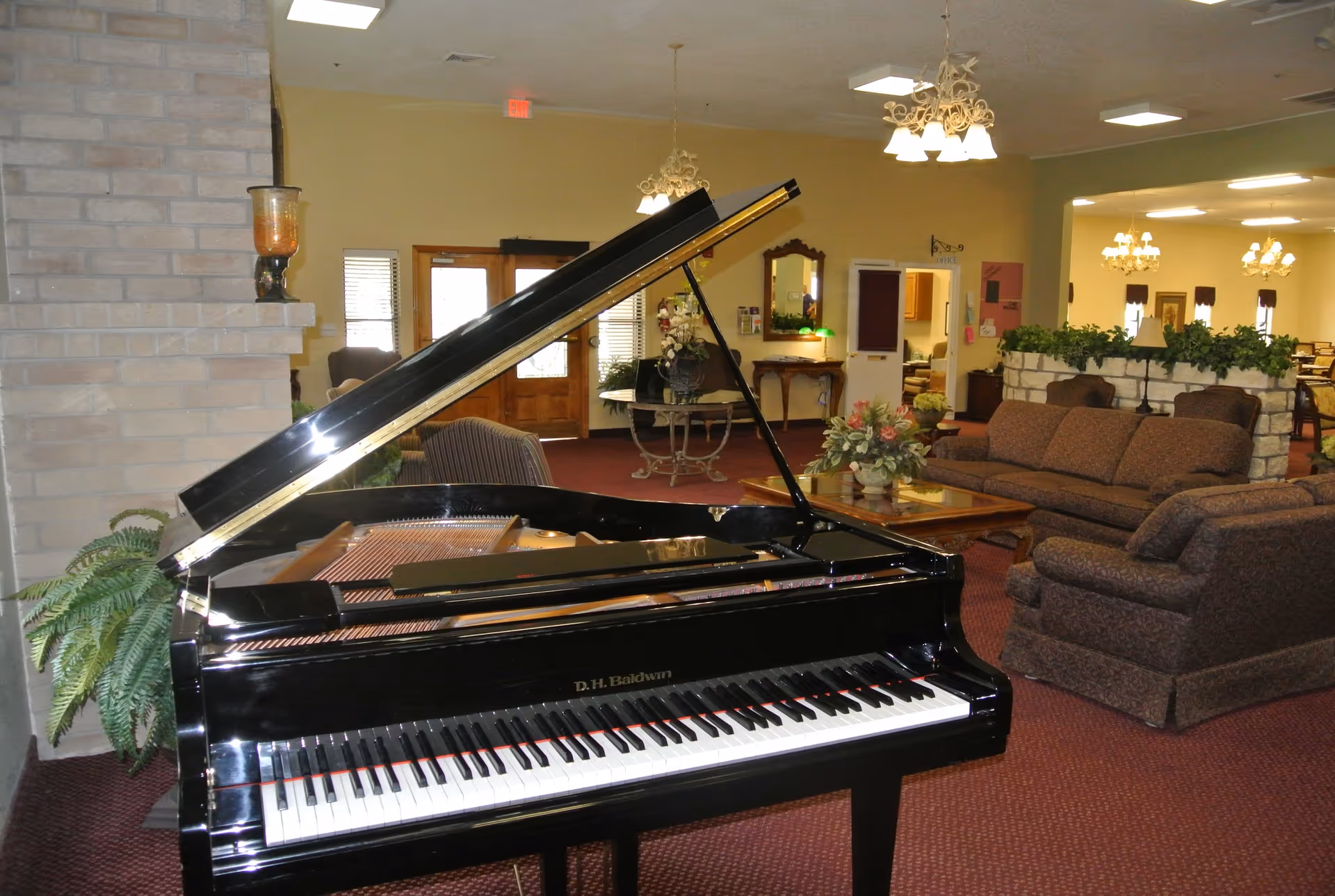 Interior of a retirement community common area featuring a black grand piano in the foreground, a brick fireplace to the left, and a seating area with brown upholstered sofas and armchairs. The room has carpeted floors, chandeliers, and a table with a floral arrangement. There are doors and windows in the background letting in natural light.