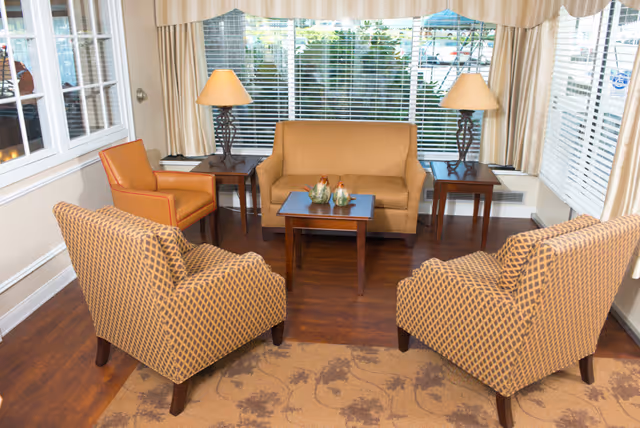 A cozy living room area with a small brown sofa, two patterned armchairs, and an orange armchair arranged around a wooden coffee table. Two side tables with matching lamps flank the sofa. Large windows with blinds and cream-colored curtains let in natural light.
