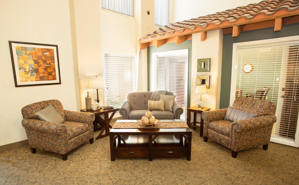 A cozy living room area with two patterned armchairs and a gray loveseat arranged around a wooden coffee table with decorative items. The room has beige walls, carpeted floor, two side tables with lamps, and large windows with blinds. There is a framed abstract artwork on the wall and a section of the wall designed to look like an exterior with a tiled roof.