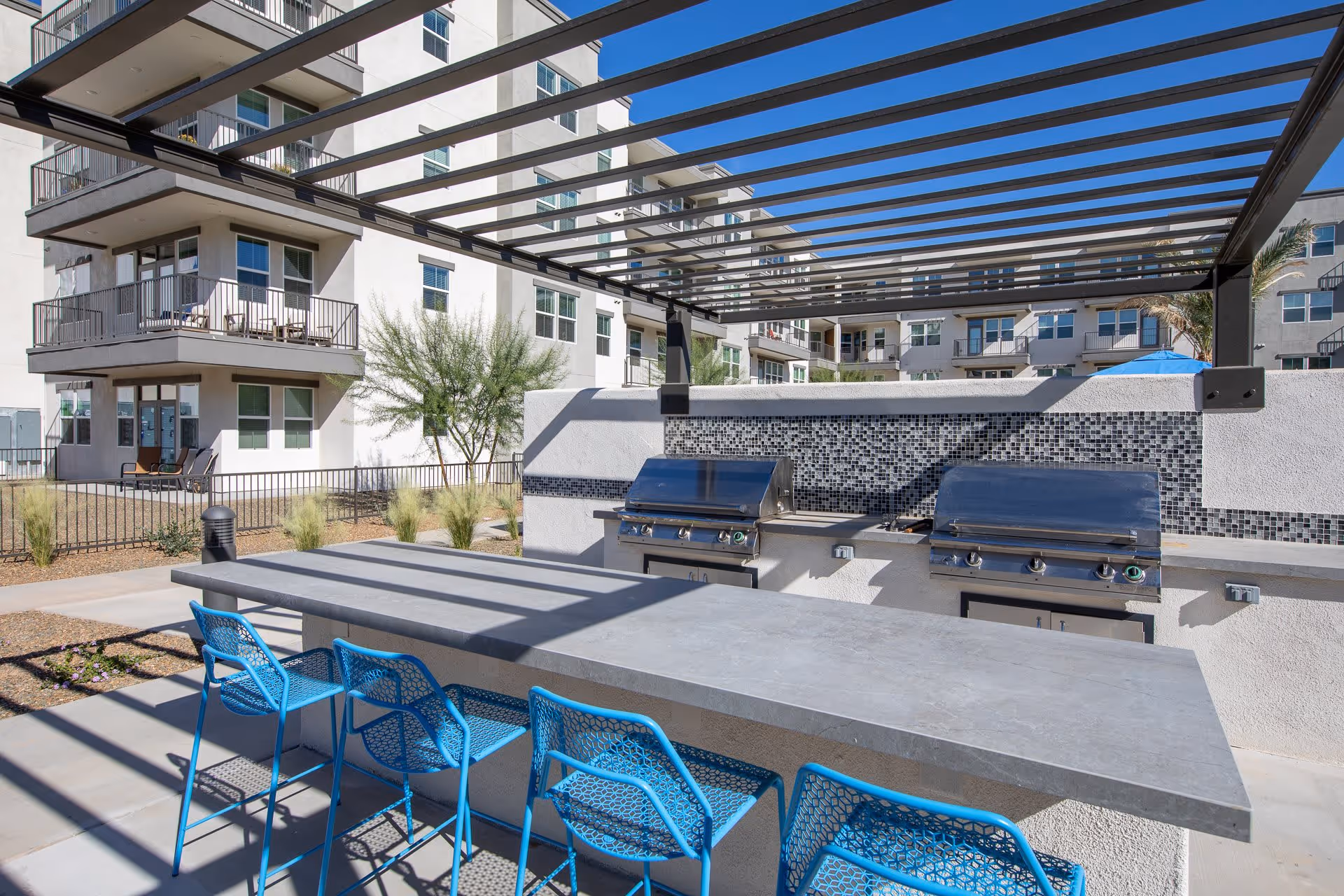 Outdoor communal grilling and seating area with blue bar stools and two stainless steel grills under a pergola in an apartment courtyard.