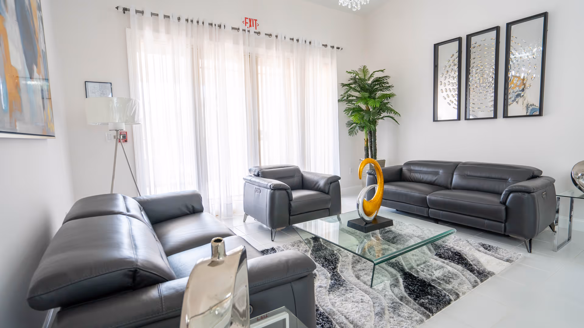 A modern living room with two black leather sofas and one black leather armchair arranged around a glass coffee table with a decorative yellow sculpture. The room has white walls, a large window with sheer white curtains, a tall green plant in the corner, a floor lamp, and three abstract framed artworks on the wall.