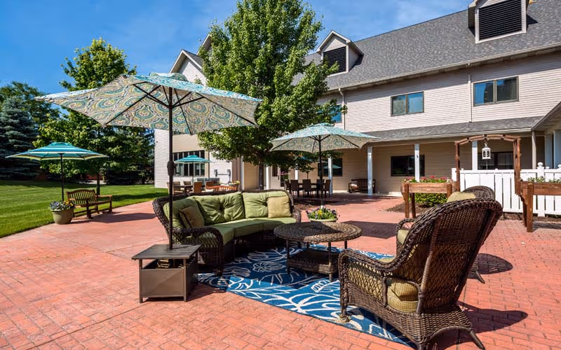 Outdoor patio area at Delta Retirement Center with wicker furniture including a green cushioned sofa, armchair, and round coffee table on a blue patterned rug. Several large umbrellas provide shade. The patio is paved with red bricks and surrounded by green grass and trees. The building in the background is beige with multiple windows and a covered porch area.