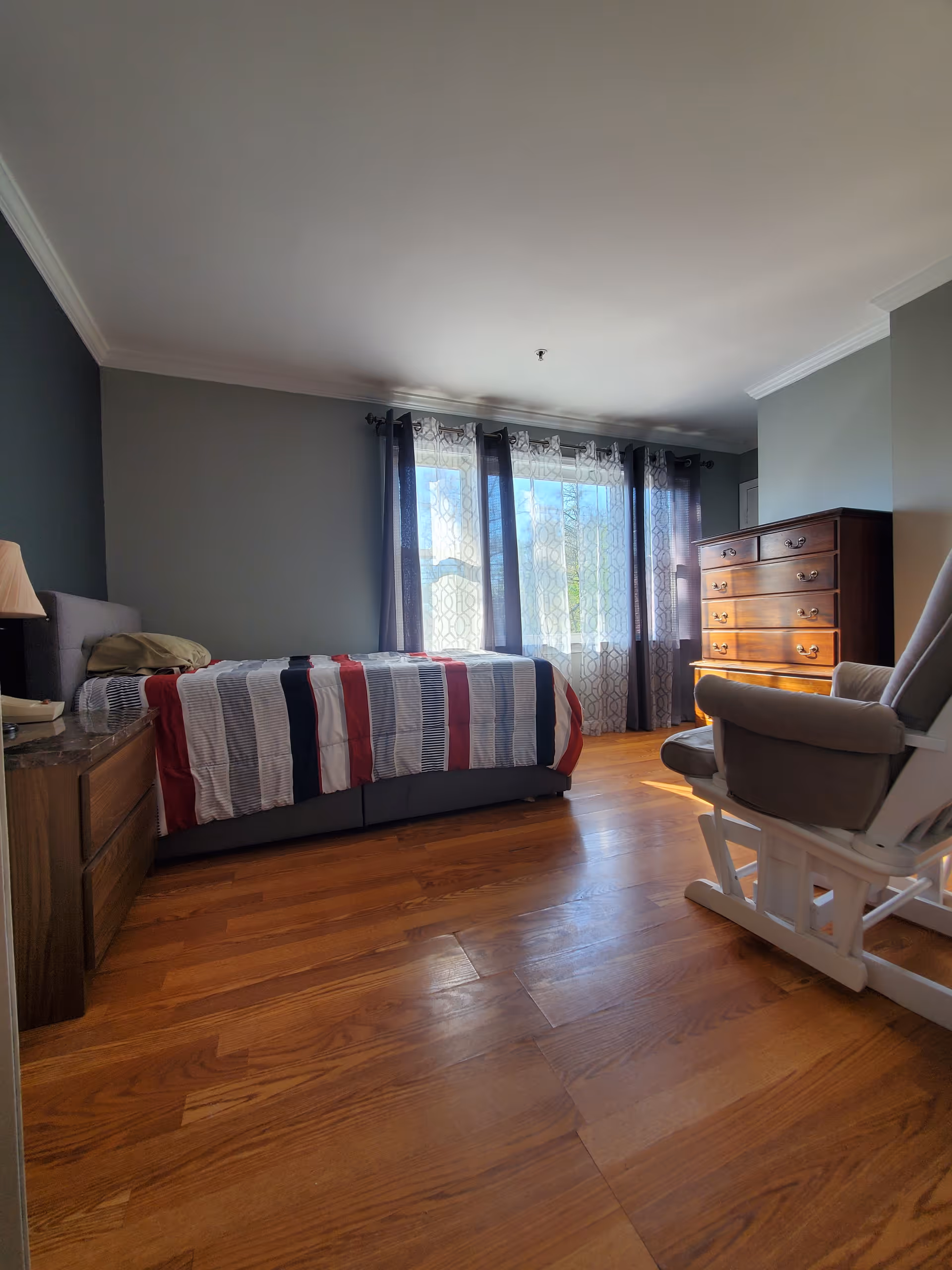 A bedroom with wooden flooring, a bed with a striped red, white, and blue comforter, a wooden nightstand with a lamp, a wooden dresser, and a cushioned rocking chair. The room has large windows with sheer and dark curtains allowing natural light to enter.