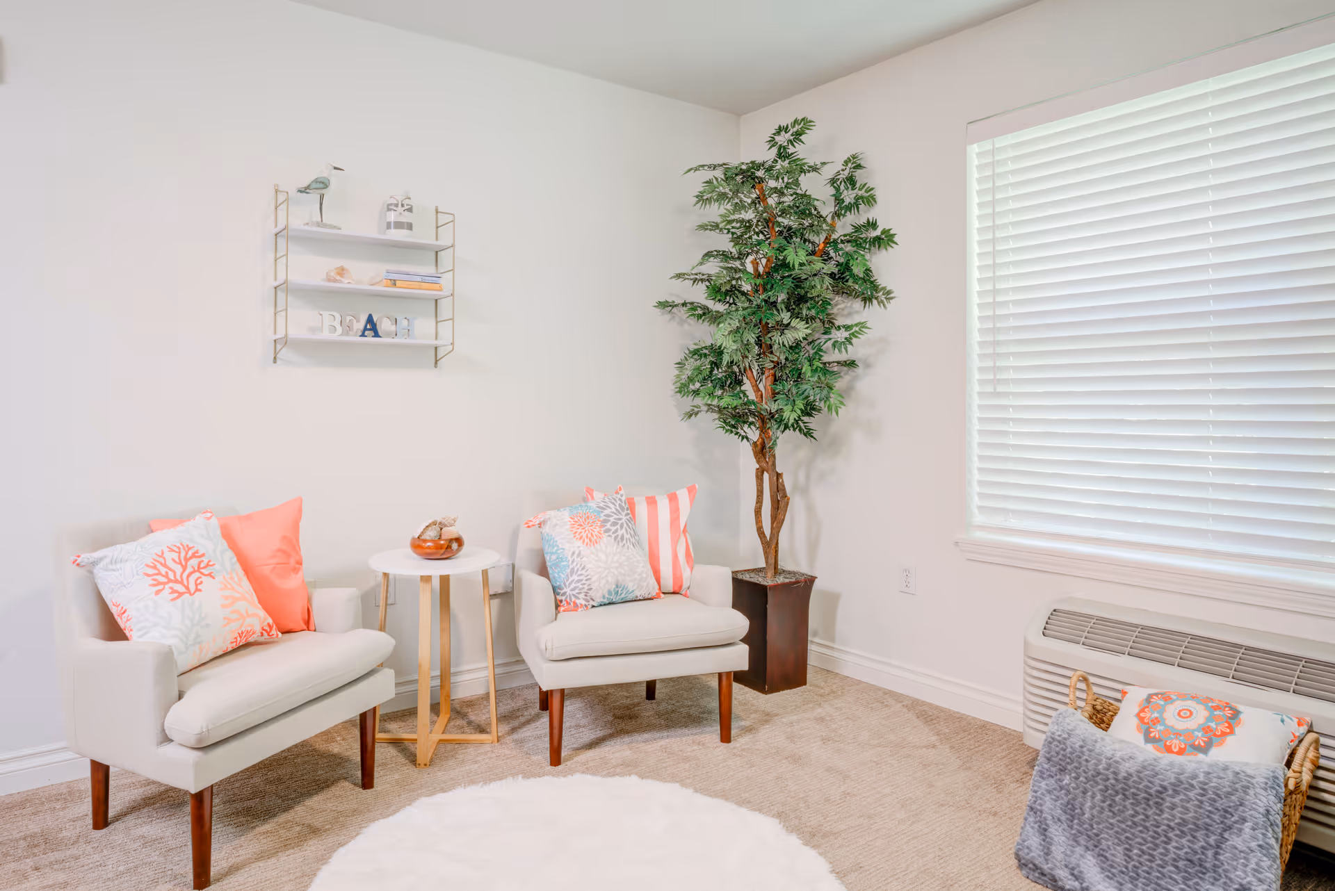 A cozy sitting area with two beige armchairs adorned with coral and patterned pillows, a small round white table between them, a tall potted artificial plant in the corner, a wall-mounted shelf with decorative items, a window with white blinds, and a basket with a blanket and pillow on the floor.