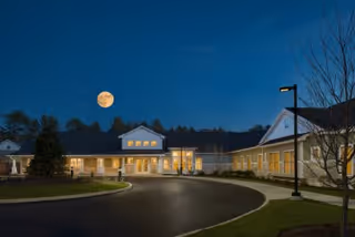 Night view of the front of a senior living facility with illuminated windows, a circular driveway, lamp posts, and a bright moon in the sky.