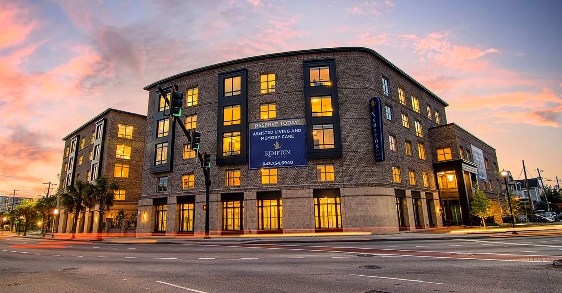 Exterior view of The Kempton of Charleston building at dusk with illuminated windows and a colorful sky. The building is a multi-story brick structure located at a street intersection with traffic lights and palm trees nearby. A large sign on the building advertises assisted living and memory care services.