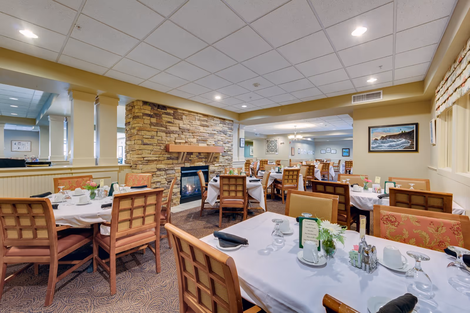 A dining room in a senior living facility with multiple tables covered in white tablecloths, set with glasses, cups, and napkins. The room features wooden chairs with cushioned seats, a stone fireplace, and framed artwork on the walls. The space is well-lit with ceiling lights and has a warm, inviting atmosphere.