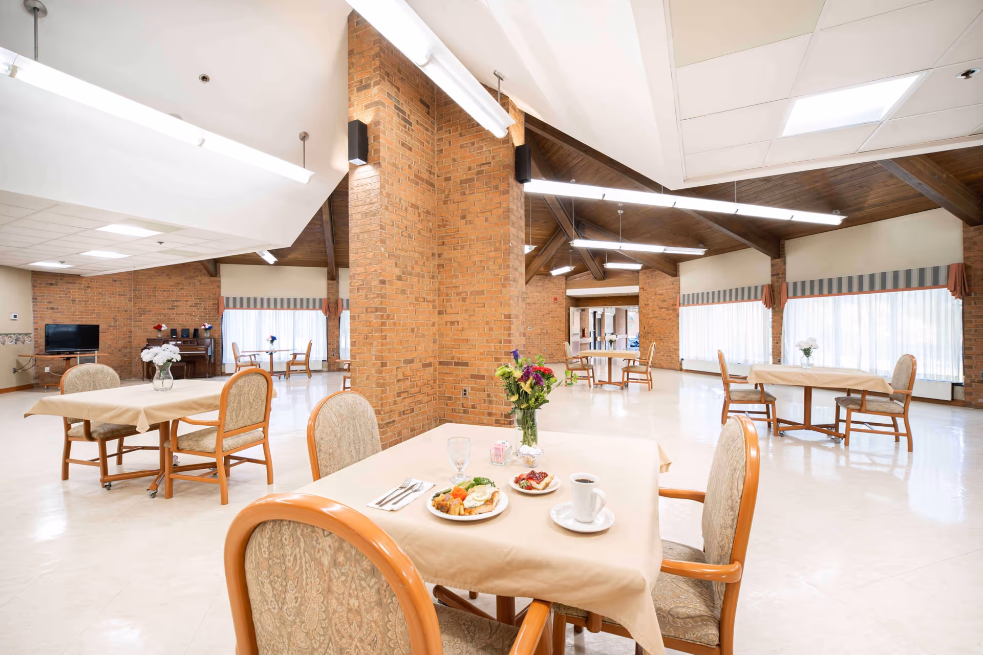A spacious dining room with several tables covered in beige tablecloths, each surrounded by cushioned wooden chairs. One table in the foreground has a plate of food, a cup of coffee, and a vase with flowers. The room features brick pillars, large windows with striped valances, and a high ceiling with exposed wooden beams and fluorescent lighting.