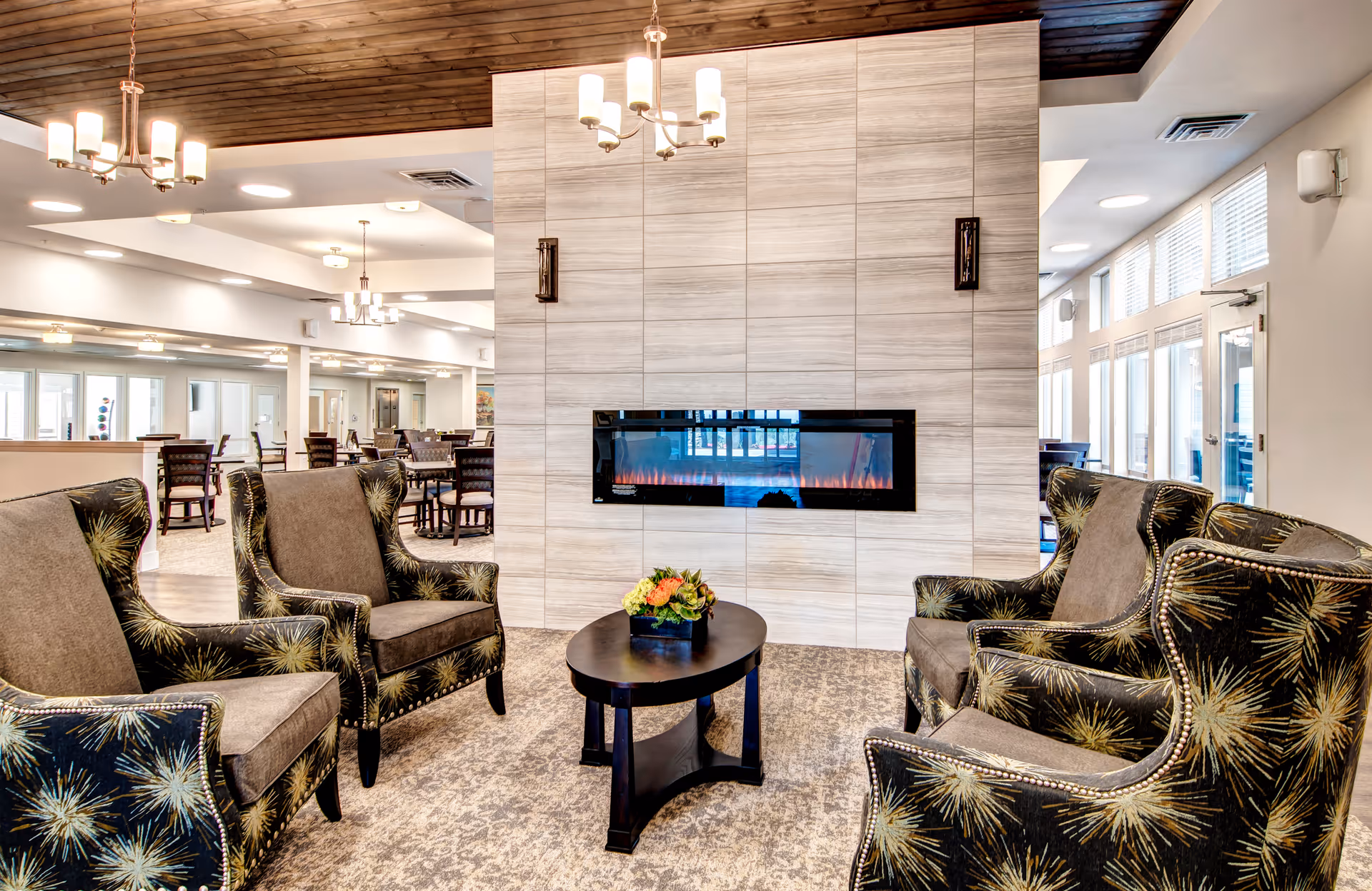 A cozy senior living community lounge area with four patterned armchairs arranged around a small black coffee table with a flower arrangement. A modern electric fireplace is mounted on a tiled wall in the background. The space features a wooden ceiling, multiple chandeliers, and large windows letting in natural light. Dining tables and chairs are visible in the background.