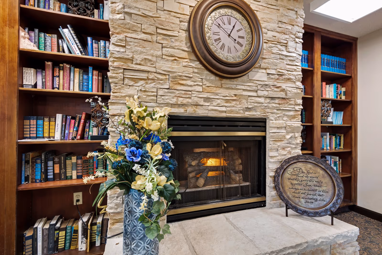 A cozy indoor scene featuring a stone fireplace with a fire burning inside. Above the fireplace is a large round clock with Roman numerals. On the left side of the fireplace is a wooden bookshelf filled with books, and on the right side is another bookshelf with more books. In front of the fireplace is a decorative vase with yellow and blue flowers, and a round plaque with an inspirational quote is displayed on the hearth.