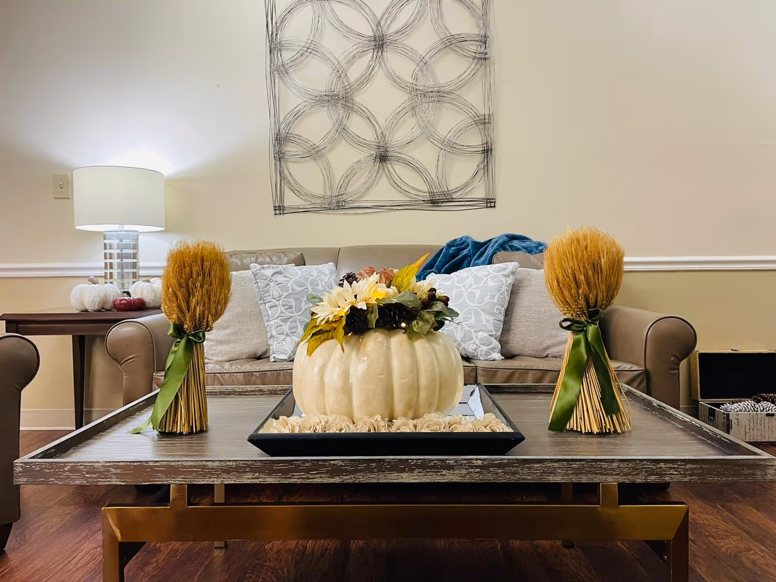 A cozy living room area with a brown leather sofa adorned with patterned pillows and a blue throw blanket. In front of the sofa is a wooden coffee table with a decorative white pumpkin centerpiece surrounded by flowers and two bundles of wheat tied with green ribbons. A side table with a modern lamp and small decorative pumpkins is visible to the left. A wall decoration featuring overlapping circles hangs above the sofa.
